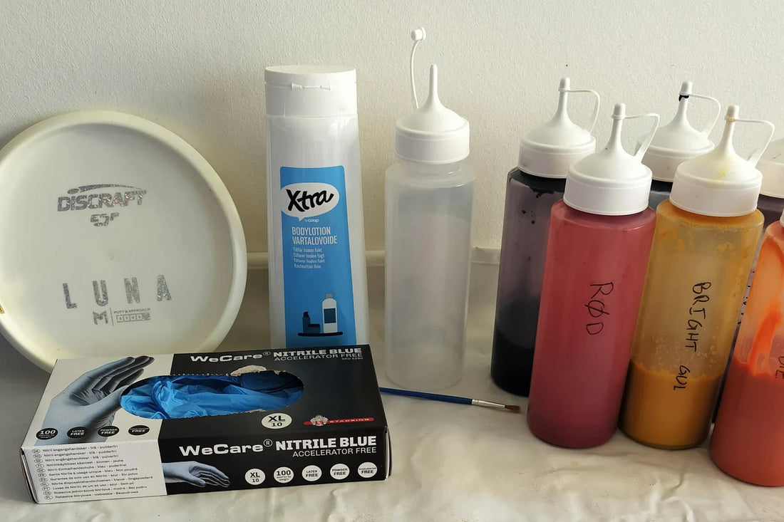 Utrustning för disc dye: en vit disc, flaskor med färg, handskar och en pensel