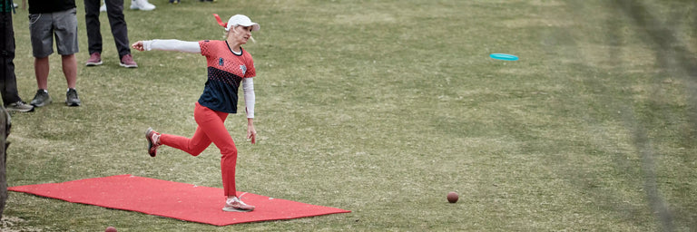 En kvindelig DGA sponsoreret disc golfer står driver sin disc fra en rød teepad med tilskuere bag hende